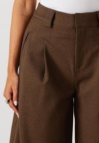 Bruine geplooide broek met een soepele textuur, voorzien van zijzakken en een op maat gemaakt tailleband. Close-up weergave die de stofdetails laat zien.