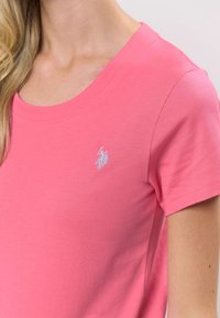 Ružové bavlnené tričko s okrúhlym výstrihom a krátkymi rukávmi. Na hrudi má malú vyšívanú značku. Jemná textúra.