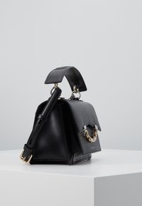 Borsa in pelle nera con manico superiore, dettaglio in catena dorata sulla patta e tracolla regolabile, appoggiata su una superficie bianca.