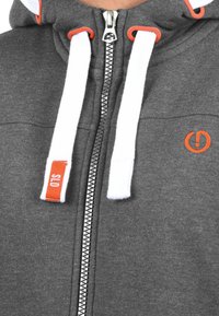 Haut zip gris avec cordons blancs, accents orange et un logo. Présente une fermeture éclair en métal et une texture de tissu douce.