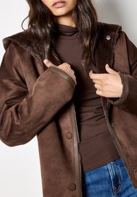 Giacca di suede marrone con cappuccio foderato in pelliccia sintetica, bottoni a pressione e maniche texture. Indossata sopra un dolcevita marrone e jeans blu.