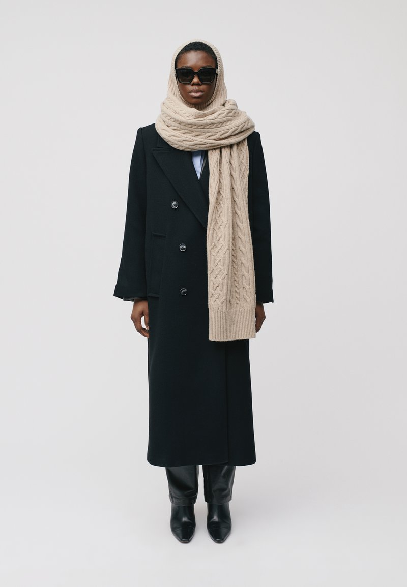 Manteau noir à double boutonnage avec de grands boutons, superposé sur une chemise bleu clair, accompagné d'une écharpe en tricot crémeuse et épaisse et d'un pantalon noir ajusté.