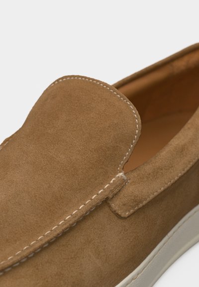 Gros plan sur une chaussure en daim marron à enfiler avec des coutures blanches et une semelle en caoutchouc beige.