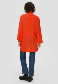 Übergroßer oranger Strickpullover mit hohem Ausschnitt und überschnittenen Schultern, kombiniert mit dunkelblauen geraden Jeans und schwarzen Stiefeletten.