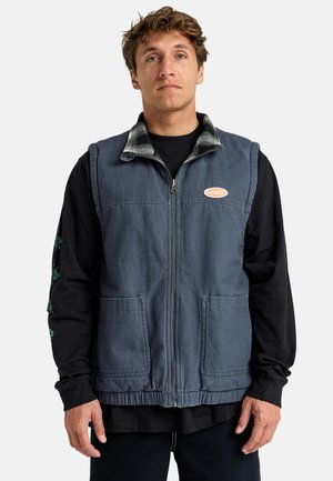 Denim vest in dark blue with a zip front, two front pockets, and a plaid collar. Worn over a black long-sleeve shirt.