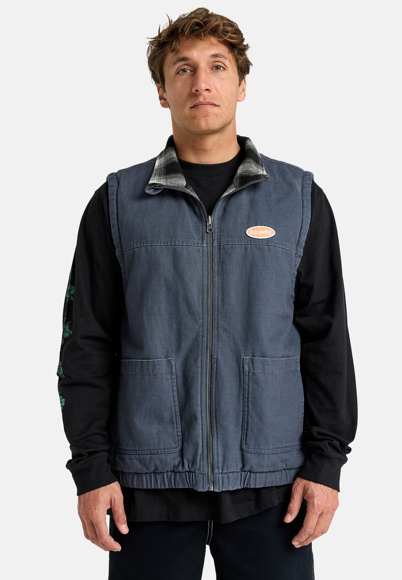 Denim vest in dark blue with a zip front, two front pockets, and a plaid collar. Worn over a black long-sleeve shirt.