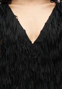 Μαύρο φόρεμα με βαθύ V-λαιμόκοψη και υφασμάτινη υφή, διακοσμημένο με μακριές, κάθετες φουντίτσες σε όλο το μήκος.