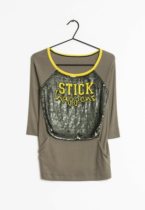 Haut à manches longues gris avec bordure jaune et panneau avant noir pailleté affichant la phrase « STICK happens » en lettres jaunes, suspendu sur un cintre noir.