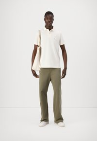 Kerja polo berwarna krim dengan kerah, celana lebar khaki, dan sepatu sneakers putih. Model membawa tas jinjing beige. Latar belakang polos.