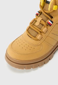 Botas de senderismo de color marrón claro con parte superior de ante texturizado, detalles de malla y forro sintético amarillo. Cuenta con cordones multicolores y suela de goma robusta.