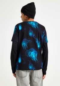 Sort t-shirt med blåt medusa-print, korte ærmer, og et sort langærmet lag underneath, parret med grå jeans.