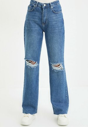 Blaue Straight-Leg-Jeans mit kaputten Knien, getragen mit weißen Sneakern und einem hellen, in die Hose gesteckten Oberteil.