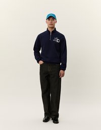 Pullover navy a zip con logo bianco, abbinato a pantaloni in denim scuro e scarpe nere. Modello che indossa un cappellino azzurro brillante. Sfondo semplice.
