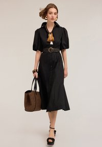 Abito nero a lunghezza ginocchio con maniche a sbuffo, chiusura con bottoni, vita cinchata e una cintura larga. Gli accessori includono una borsa intrecciata e sandali con tacco.