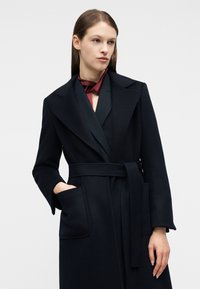 Γυναίκα που φοράει σκούρο μπλε παλτό με ζώνη, μεγάλους γιακάδες και τσέπες-μπαλώματα, συνδυασμένο με μπουρνούζι σε μπορντό χρώμα και σκούρο εσωτερικό ένδυμα.