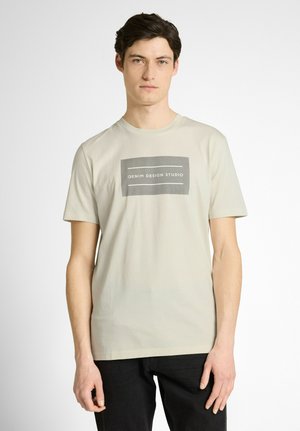 Junger Mann trägt ein beiges T-Shirt mit einem grauen rechteckigen Logo, auf dem "Denim Design Studio" steht, kombiniert mit schwarzen Hosen.