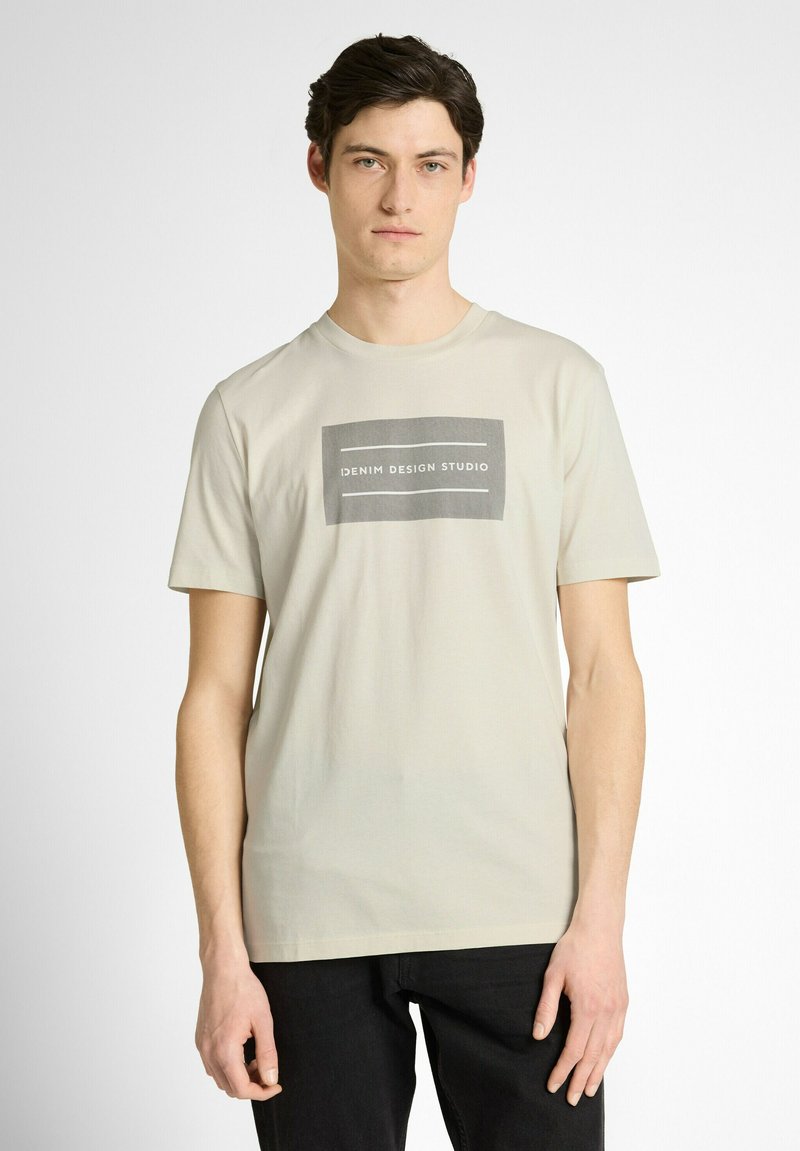 Jeune homme portant un t-shirt beige avec un logo rectangulaire gris indiquant "Denim Design Studio", associé à un pantalon noir.