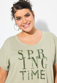 Camiseta de manga corta verde claro con la inscripción "PRIMAVERA" impresa en brillantes lentejuelas verdes. Tejido suave con un ajuste holgado y escote ancho.