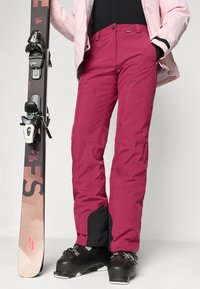 Fukszia színű sínadrág, laza szabással, megerősített fekete panelokkal, cipzáras zsebekkel és tartós anyagból készült. Síléc és cipő kíséretében.