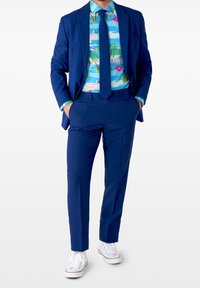 Abito blu navy dal taglio aderente, abbinato a una camicia con motivo tropicale multicolore con fenicotteri e sneaker bianche.