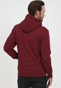 Felpa maroon con cappuccio regolabile con cordino, polsini e orlo a costine. Presenta una texture di tessuto liscia, mostrata dal retro.