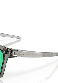 Occhiali da sole con montatura in plastica grigia, lenti verdi e braccia trasparenti testurizzate. Presentano dettagli di marca e un design angolare elegante.