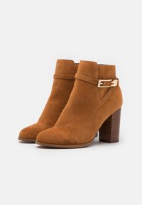 Botas de tobillo de ante marrón con una ligera curvatura en la parte superior, que presentan un acento de hebilla dorada y un tacón de madera apilado.