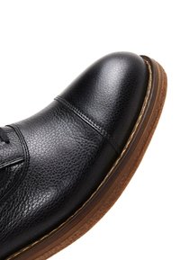 Scarpa in pelle nera con punta rotonda, tomaia con texture e suola in gomma marrone. Presenta una cucitura lungo il davanti.