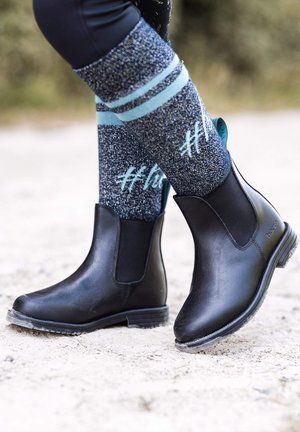Schwarze Leder-Ankle-Boots mit elastischen Seitenpaneelen und strukturierten Sohlen, kombiniert mit blauen und grauen gemusterten Socken mit Streifen und Hashtags.