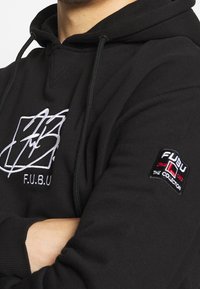 Černá mikina s kapucí a šnůrkou, na přední straně vyšívané bílé logo a na rukávu nášivka s nápisy "FUBU" a "The Collection."