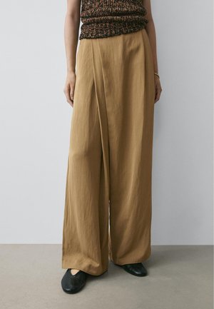 Donna che indossa pantaloni color cammello a vita alta e gamba larga, un top marrone lavorato a maglia senza maniche, ballerine nere e un braccialetto d'oro al polso sinistro.