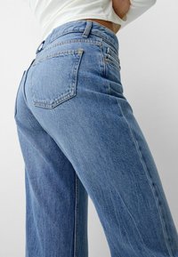 Närbild av en person som bär ljusblå högmidjade denim jeans och en vit kortärmad topp med lång ärm mot en enkel vit bakgrund.