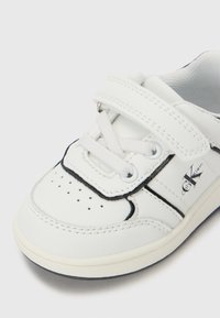 Baskets blanches pour enfants avec une sangle Velcro et des lacets, embout perforé, bord noir et logo Calvin Klein sur le côté.