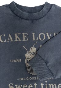 Sudadera navy con cuello acanalado, que presenta texto en dorado metálico y un gráfico de cupcake. Material suave de algodón con mangas largas.