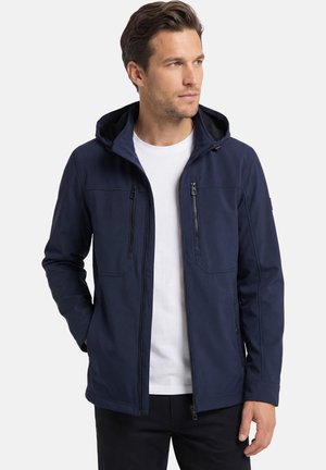 Mann trägt marineblaue Kapuzenjacke über weißem Hemd, steht mit Händen in den Taschen und blickt nach rechts vor schlichtem Hintergrund.
