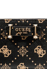 Cartera negra texturizada con patrón del logo de Guess en beige y placa metálica dorada con el texto "GUESS EST 1981".