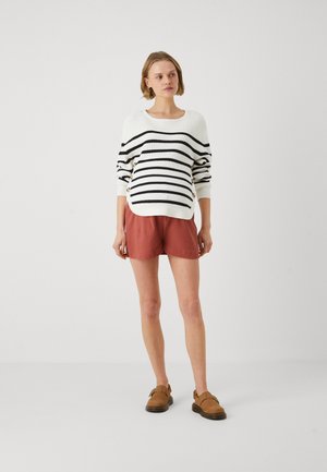 Woman wearing a white and black striped sweater with gold buttons, rust-colored shorts, and brown buckle shoes, standing against a plain wall.