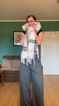 Eșarfă din amestec de lână, albă cu dungi gri, cu margini franjurate. Utilizatorul ține eșarfa aproape, purtând un top negru și pantaloni largi gri.