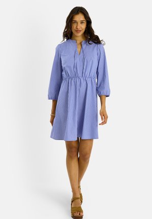 Mujer con un vestido de rayas verticales azules y blancas con mangas tres cuartos y sandalias cuña marrones, caminando sobre un fondo blanco liso.