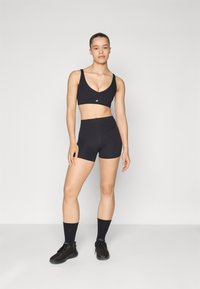 Crna sportska bralette s dubokim V-izrezom i logom, u kombinaciji s crnim šortsevima visokog struka. Oba od rastezljivog, glatkog materijala. Crne tenisice i čarape.