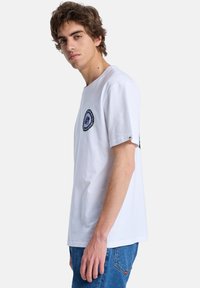 Bílé bavlněné tričko s kulatou grafickou nášivkou na levé straně hrudníku a krátkými rukávy, spojené s modrými džínovými kalhotami.