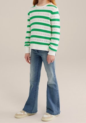 Kind trägt einen weiß-grün gestreiften Strickpullover, ausgewaschene blaue Schlagjeans und beige-weiße Turnschuhe, steht vor neutralem Hintergrund.