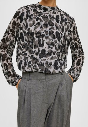 Woman wearing pleated long-sleeve leopard print blouse tucked into high-waisted gray pleated trousers with hands in pockets.