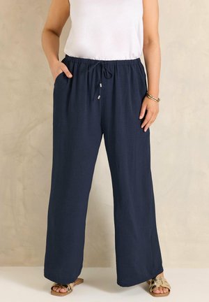 Mujer con pantalones anchos de pierna en azul marino con cordón, camiseta sin mangas blanca, sandalias doradas y pulseras, de pie con una mano en el bolsillo.