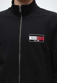 Veste noire zippée avec un col haut présentant un patch logo rectangulaire en blanc, rouge et noir avec le texte "HILFIGER Est. 1985".