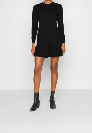 Woman wearing a black long-sleeve dress with ruffled hem and black ankle boots with metal toe caps, standing against a plain background.