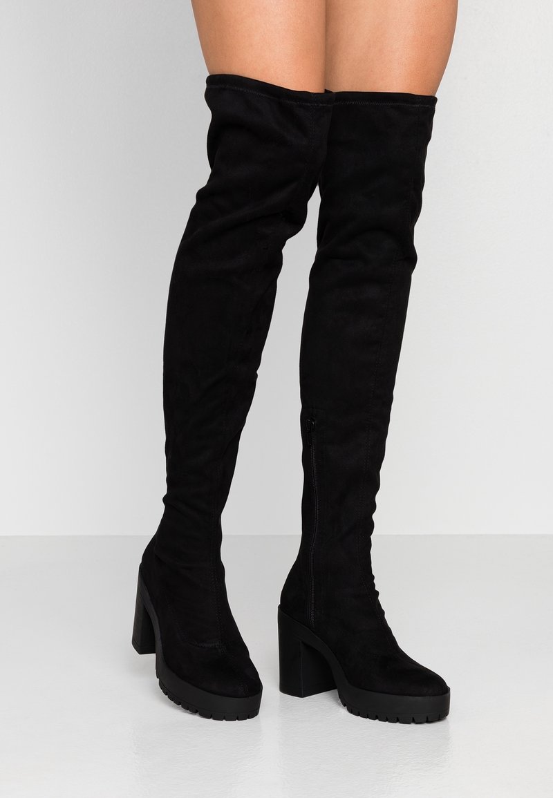 Bottes en daim noir montant jusqu'à mi-cuisse avec talons hauts et épais et fermeture éclair latérale, portées sur des jambes nues, debout sur un sol gris clair.