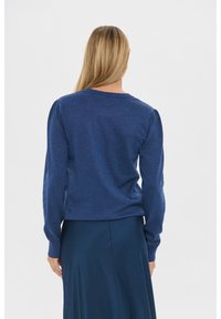 Szőke hajú nő hosszú ujjú kék pulcsiban és hozzá illő sötétkék szoknyában áll egy egyszerű háttér előtt, háttal a kamerának.