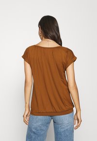 Top marrone a maniche corte con una vestibilità rilassata, caratterizzato da un colletto rotondo e un dettaglio arricciato sulla schiena, abbinato a jeans azzurri chiari.