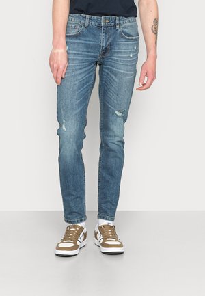 Jeans de ganga azul claro com um corte slim, apresentando áreas desgastadas e um desbotamento subtil. Combinados com ténis castanhos e brancos.